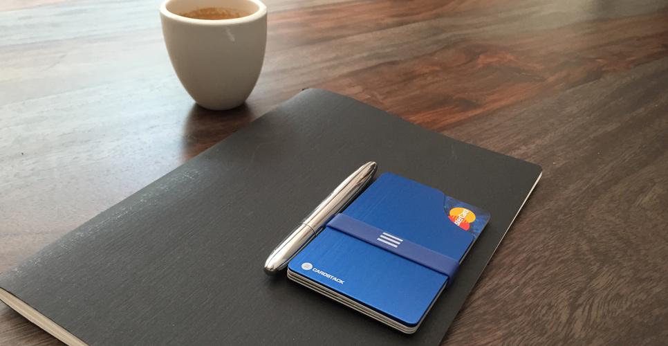 Le portefeuille Cardstack, bleu, posé sur un carnet noir à côté d’un stylo et d’un café